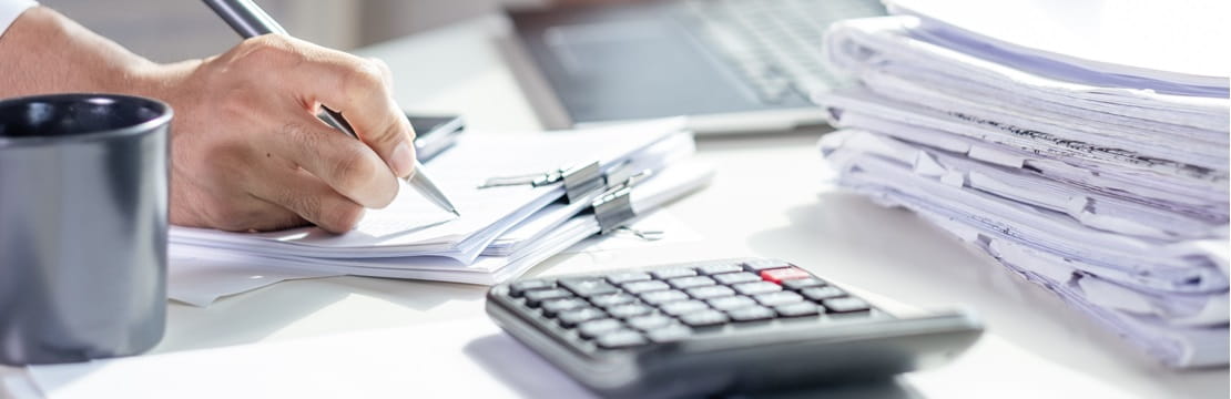 Person writing on documents at a desk with a calculator, coffee mug, laptop, and a stack of papers.